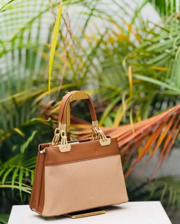 A stylish handbag with brown leather and beige fabric, adorned with gold accents, set against lush tropical greenery.