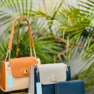 Three stylish handbags in orange, beige, and navy sit against a lush green backdrop of palm leaves.
